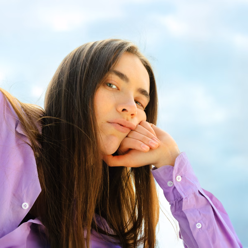 Woman with long hair resting her chin on her hands, looking thoughtful against a cloudy sky.
