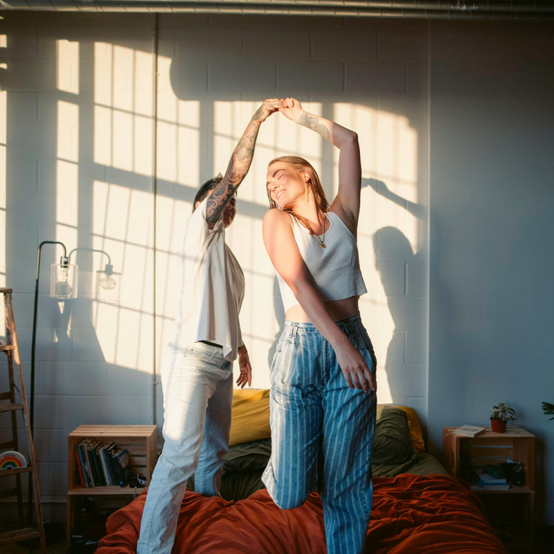 Zwei Personen stehen auf einem Bett, halten sich an den H&auml;nden und strecken die Arme nach oben