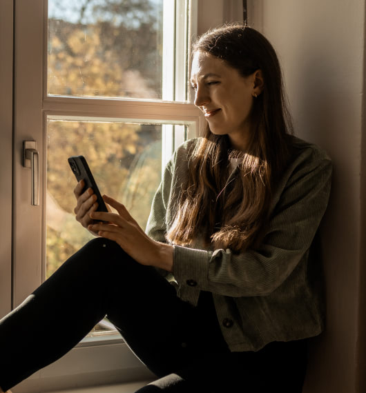 Photo : Une jeune femme est assise détendue sur un rebord de fenêtre, tenant un smartphone dans la main et souriant.